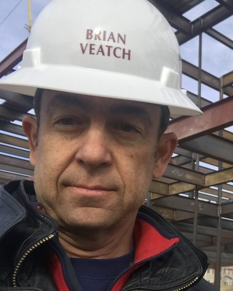 Brian Veatch, principal of Silvergate Property Management, wearing a white hard hat with his name on it at a construction site with steel framing behind him